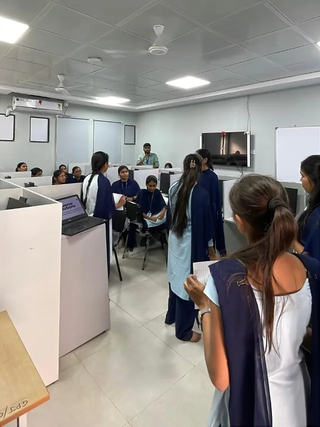 Students in navy and light blue uniforms stand and sit among white computer cubicles while a man stands at the back of a bright lab.
