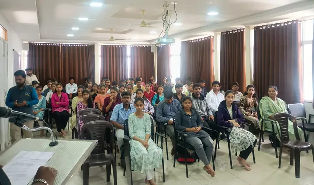 Students in rows of chairs face the front of a classroom with brown curtains, seen from behind a podium with a microphone.