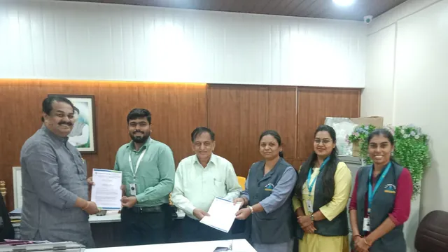 Six people in an office; two pairs hold MoU documents between them while two women in grey vests stand on the right.