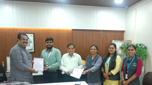 Six people stand in an office as two pairs exchange printed MoU documents.